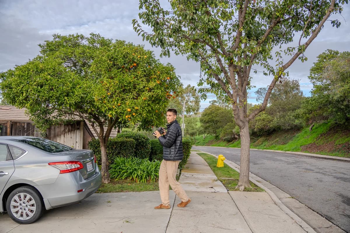 Blaine carrying his camera along a tree-lined neighborhood street